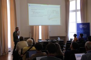 Picture of Dr Andrey Romanov, in front of the audience, standing next to the screen where his presentation is projected